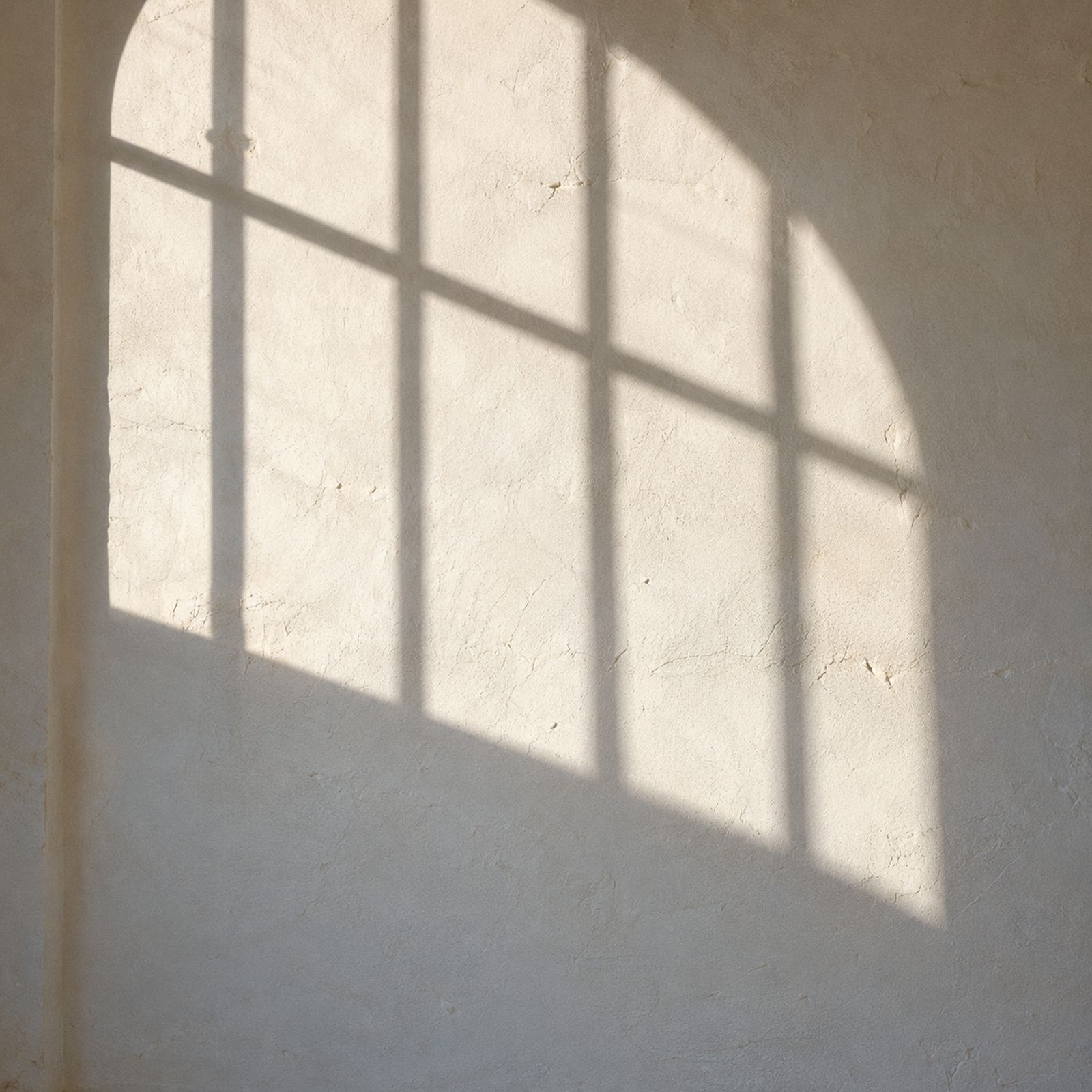Hard sun throws the shadow of an arched window grid across a lime-washed wall